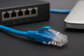 Nahaufnahme eines blauen Patchkabels mit RJ45-Stecker vor Router und Laptop