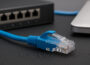 Nahaufnahme eines blauen Patchkabels mit RJ45-Stecker vor Router und Laptop